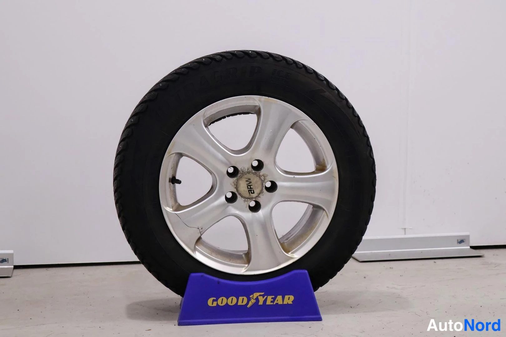 Yksityiskohta: Close-up of a single winter wheel with a silver five-spoke alloy rim and studded tire mounted on a blue Goodyear branded stand.