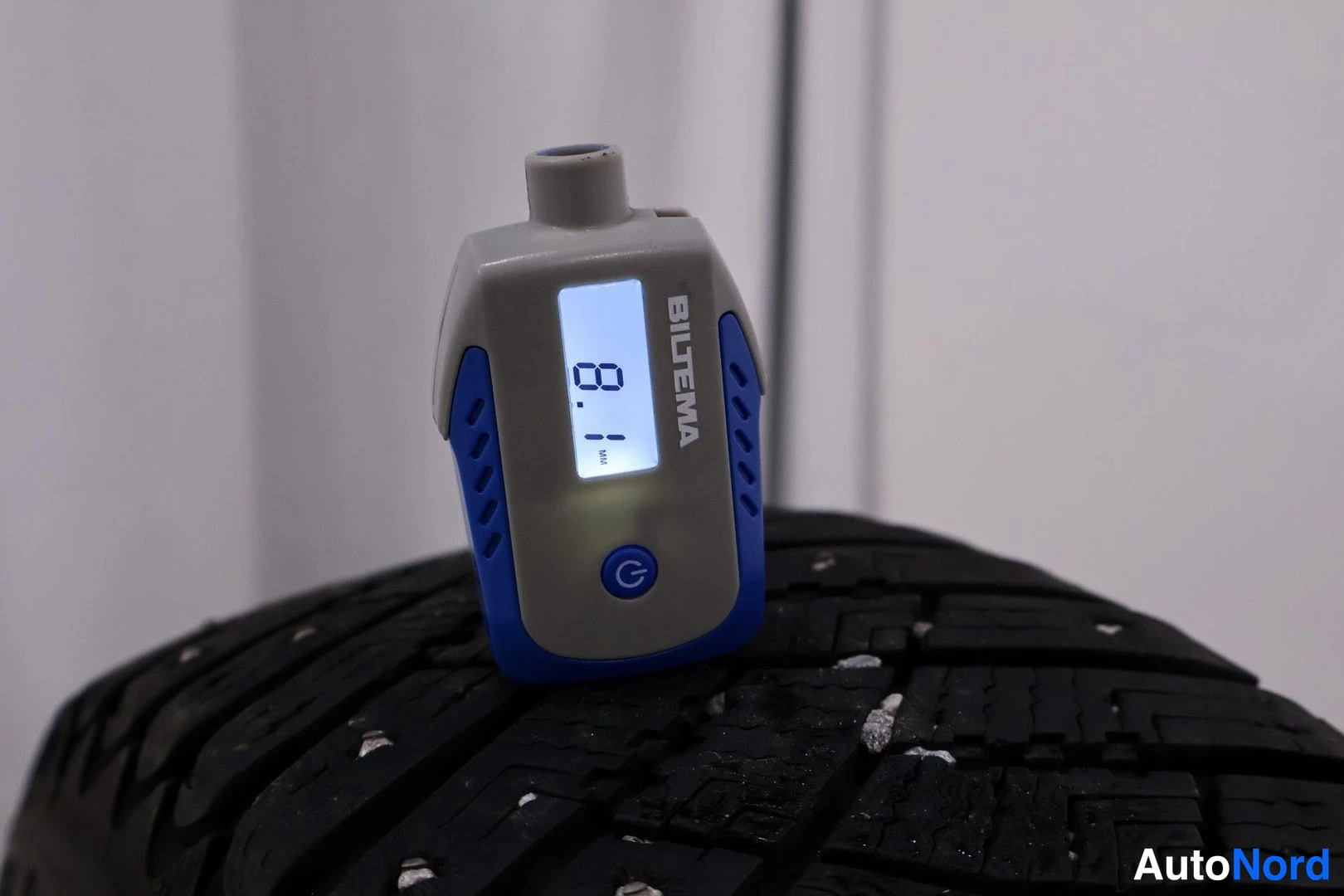 Yksityiskohta: Close-up of a Biltema digital tread depth gauge reading 8.1 mm inserted into the groove of a snow tire with embedded ice crystals.