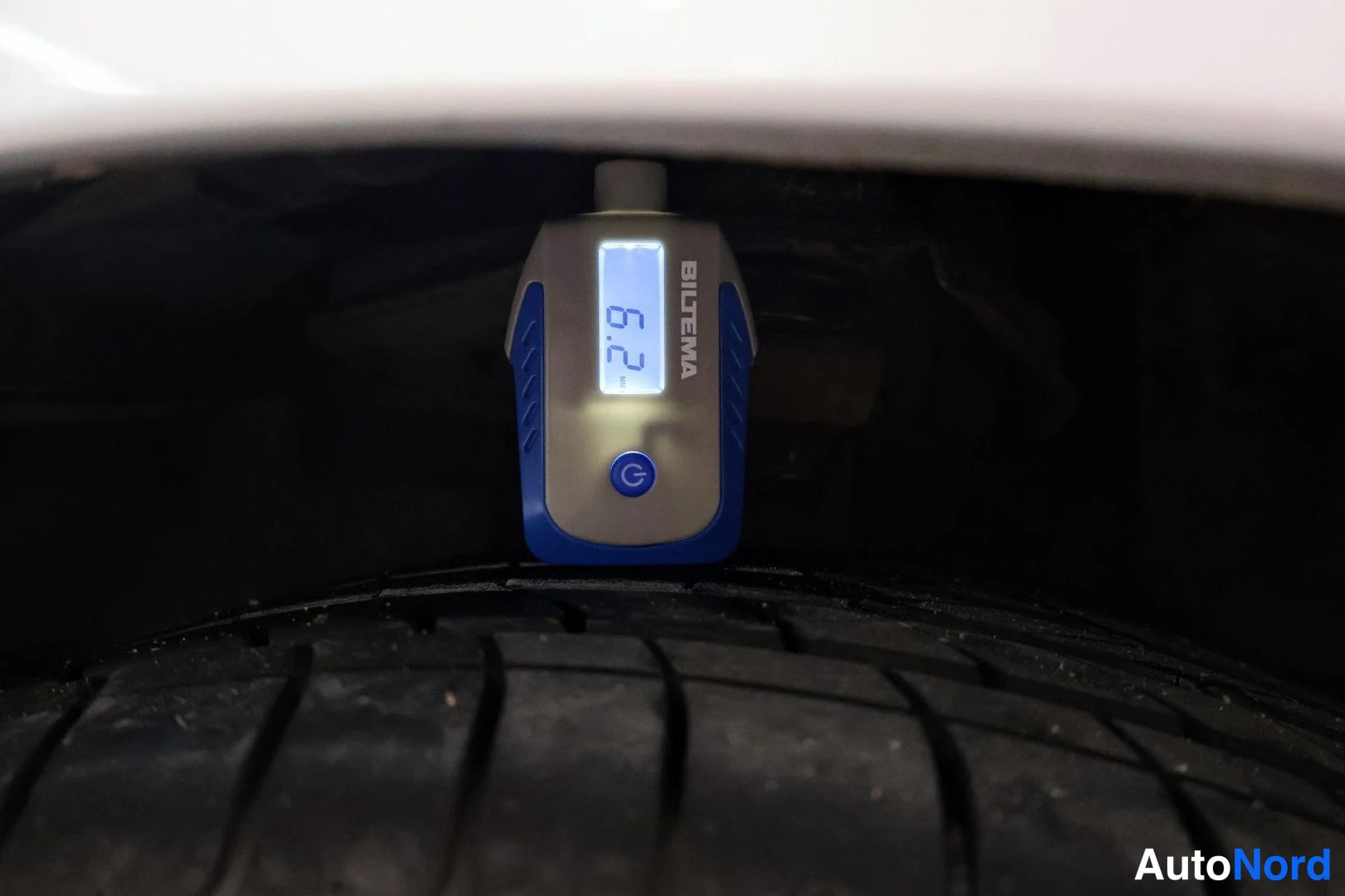 Yksityiskohta: Close-up of a Biltema digital tread depth gauge reading 6.2 mm measuring a summer tire, partially obscured by the white underbody of a vehicle.