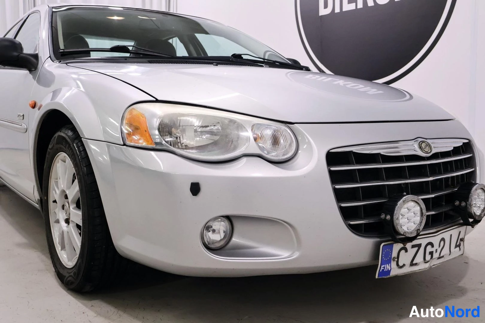 Etuviisto, nokka vasemmalle: Front three-quarter view of a white Chrysler Sebring parked in an indoor showroom, featuring a chrome grille, fog lights, and auxiliary LED lamps.