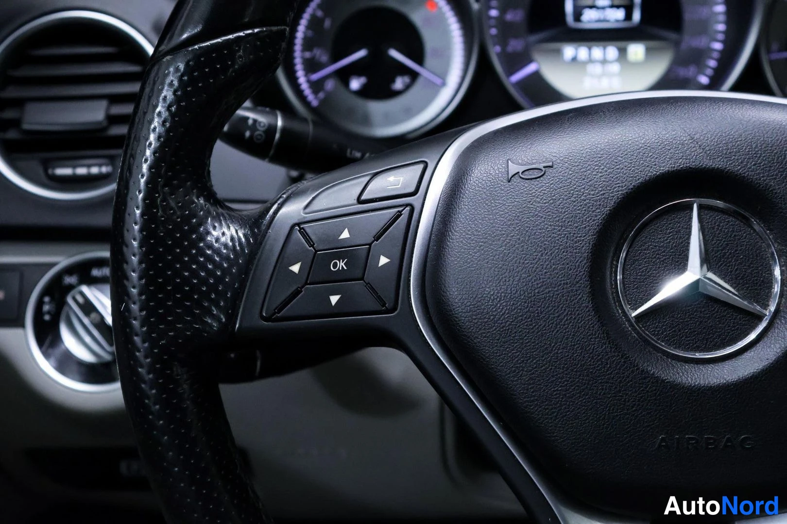Muu: Sisäkuva: Mercedes-tunnuksella varustettu ohjauspyörä ja pyörän napin painikkeet, kojelaudan mittarit taustalla.