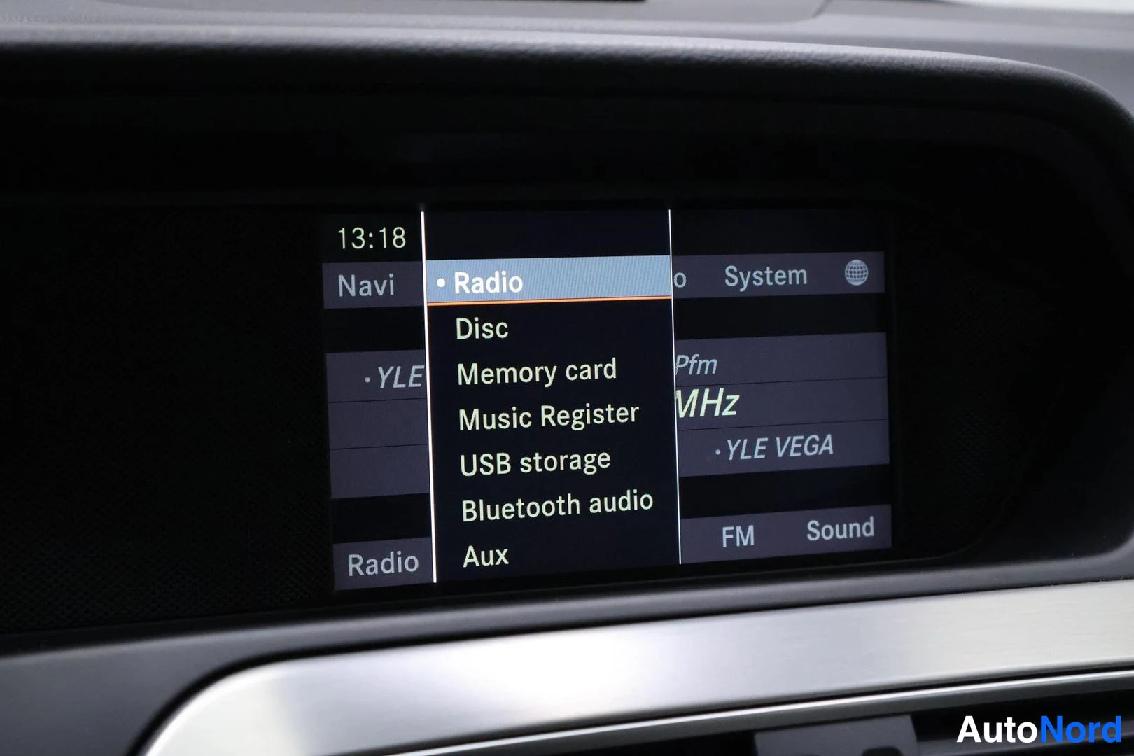 Yksityiskohta: Kojelaudan infotainment-näyttö: Radio-valikko korostettuna, vaihtoehtoina Disc, Memory card, Music Register, USB storage, Bluetooth audio ja Aux; kellonaika 13: