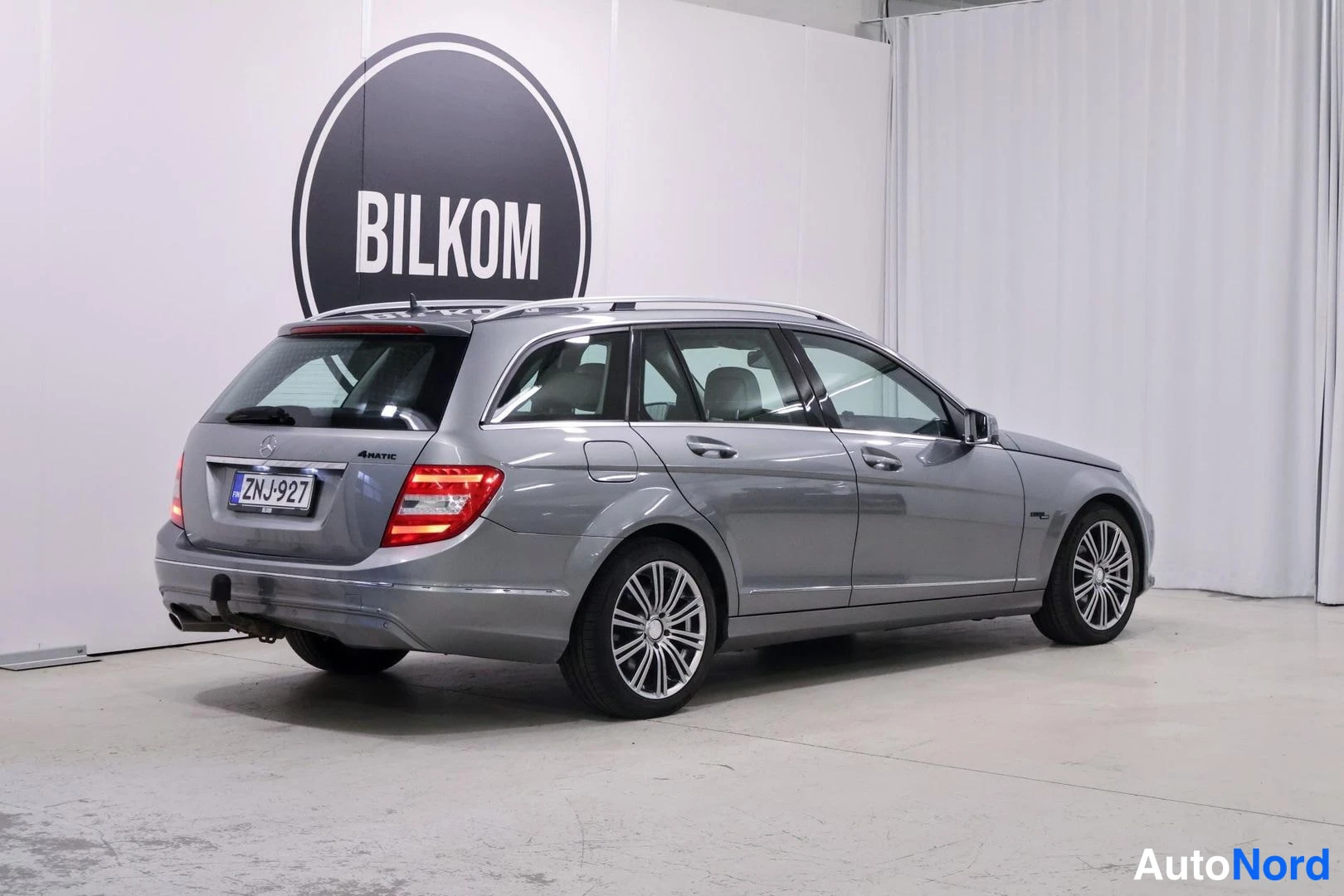 Takaviisto: Hopeanharmaa Mercedes-Benz C-farmari takaa-vinoittain kuvattuna hallissa, rekisterikilpi.