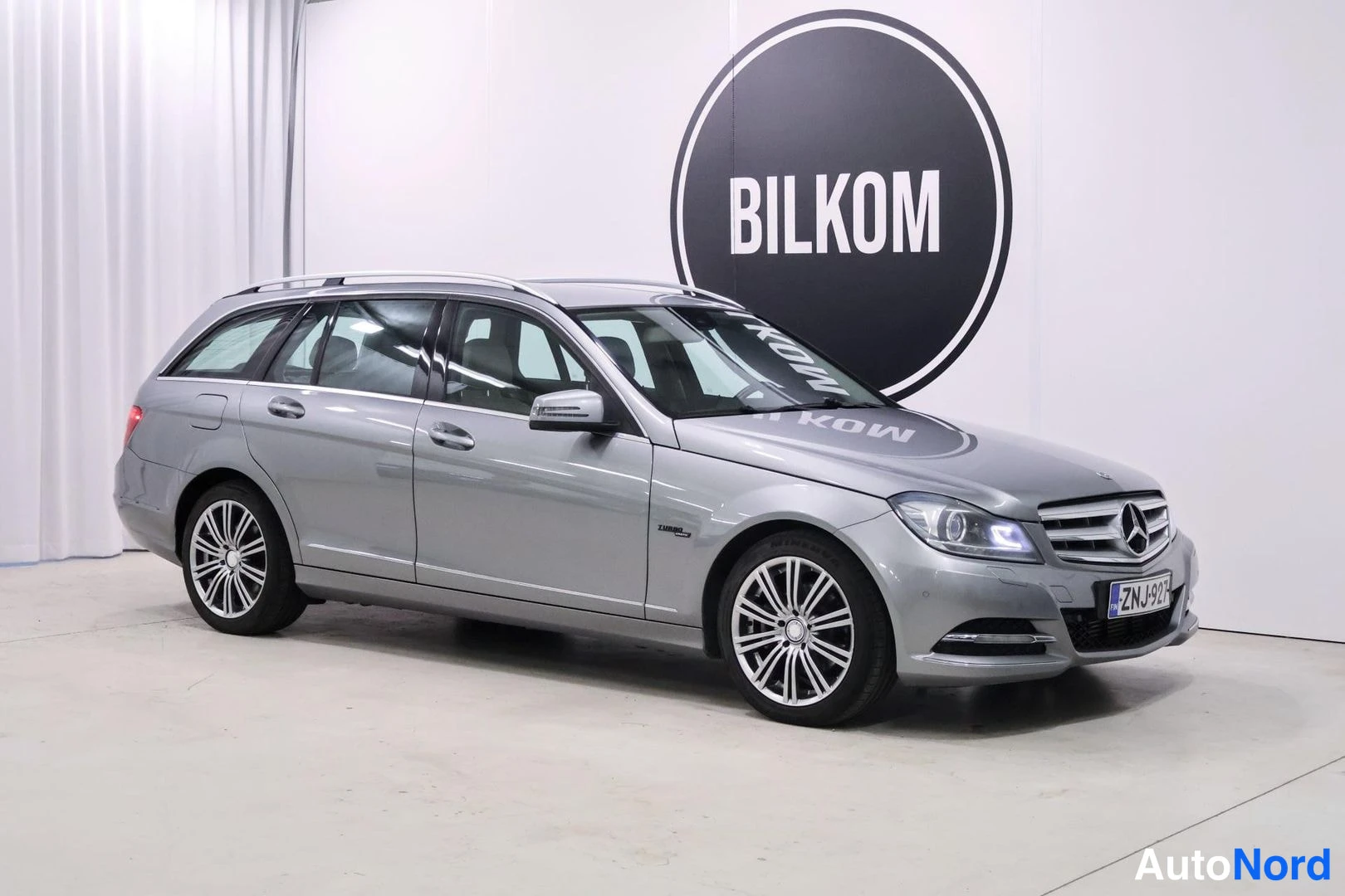 Etuviisto, nokka oikealle: Hopeanharmaa Mercedes-Benz C-farmari edestä-vinoittain kuvattuna hallissa, rekisterikilpi.