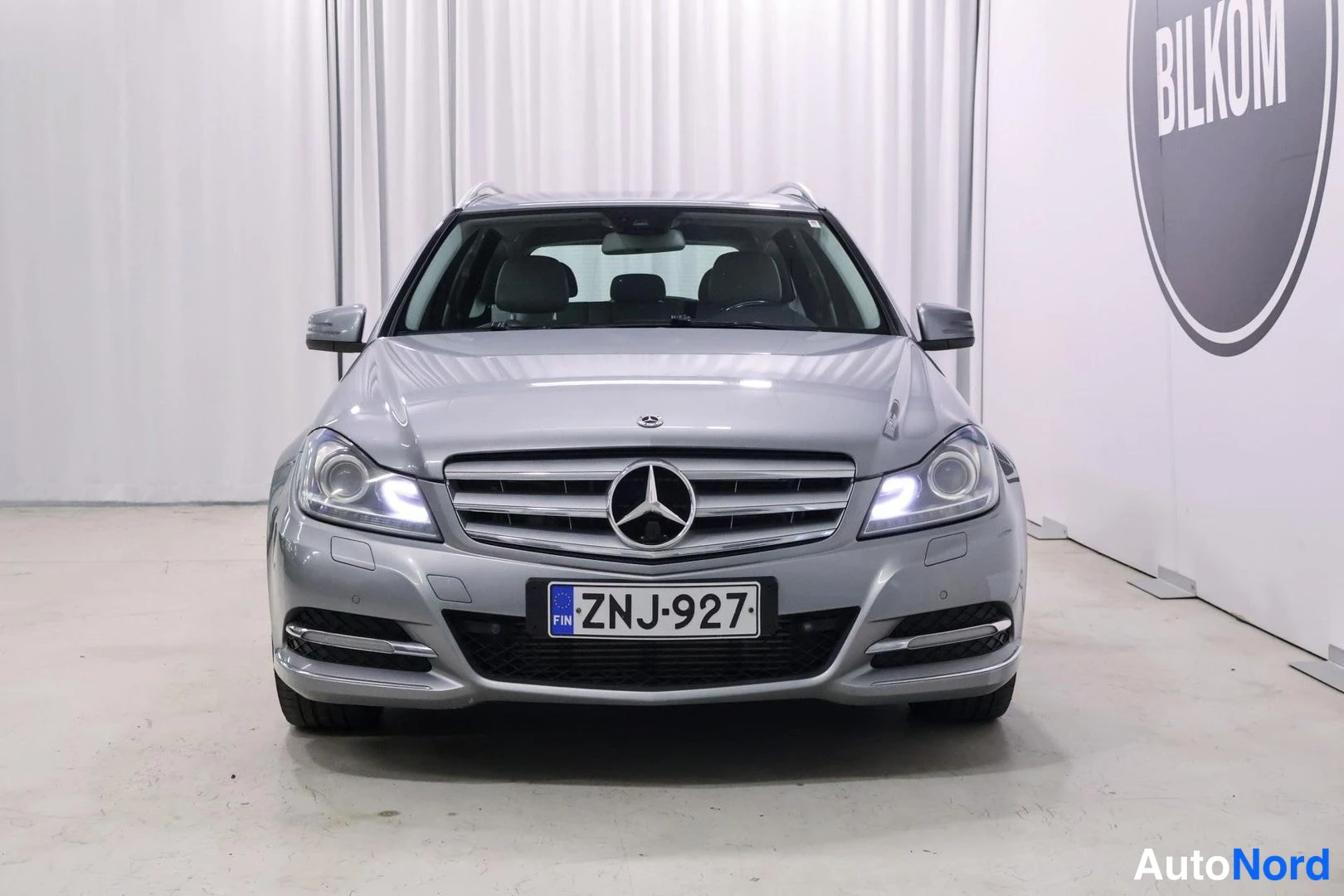 Edestä: Hopeanharmaa Mercedes-Benz C-farmari suoraan edestä kuvattuna hallissa, rekisterikilpi.