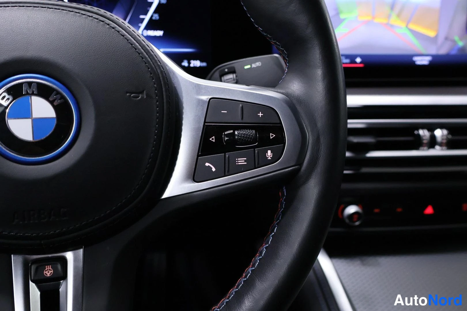 Muu: Lähikuva BMW:n ohjauspyörän oikean puhallusryhmän napeista: äänenvoimakkuus, valintaohjain, puhelin, lista ja mikrofoni. Taustalla kojelaudan näytön häivähdys.