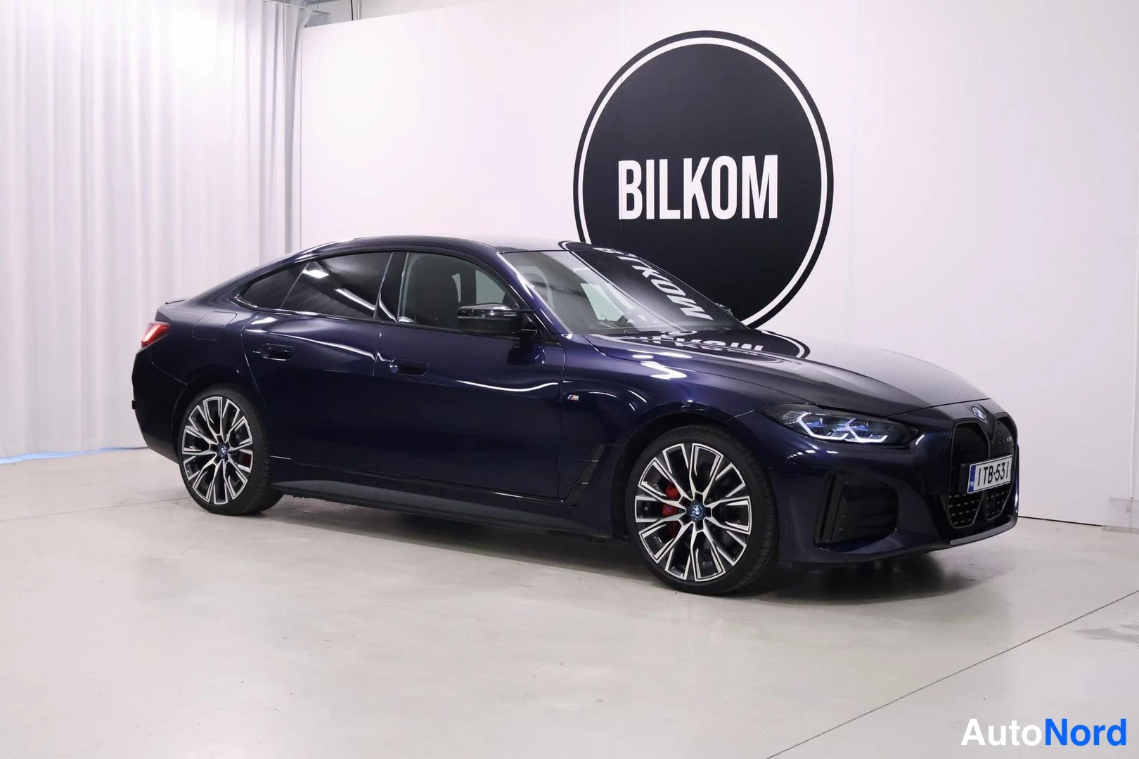 Etuviisto, nokka oikealle: Sininen BMW i4 etuviistosta vasemmalta kuvattuna sisätiloissa.