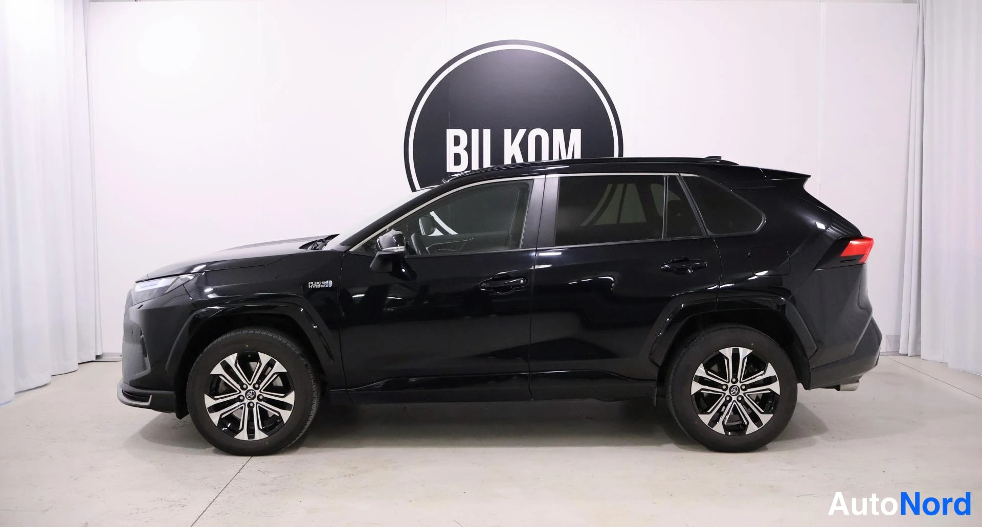 Sivulta: Musta Toyota RAV4 näkyy profiilista vasemmalta puolelta. Autoon kuuluvat kaksiväriset alumiinivanteet ja 'HYBRID'-tunnus sivussa. Taustalla valkoiset seinät ja
