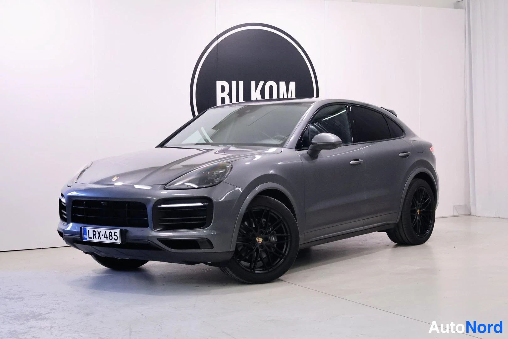 Etuviisto, nokka vasemmalle: Tummanharmaa Porsche Cayenne Coupé etukulmakuvassa sisätilassa, keula vasemmalla; mustat monipuoliset kevytmetallivanteet ja rekisterikilpi.