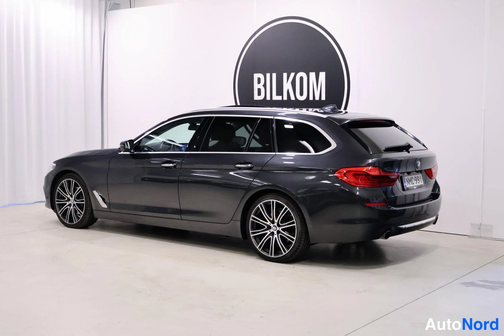 Takaviisto: Tummanharmaan BMW Touring -farmarin takaosan kolmasosakulmakuva vasemmalta, takavalot ja monitavaravanteet näkyvät valkoisessa hallissa.