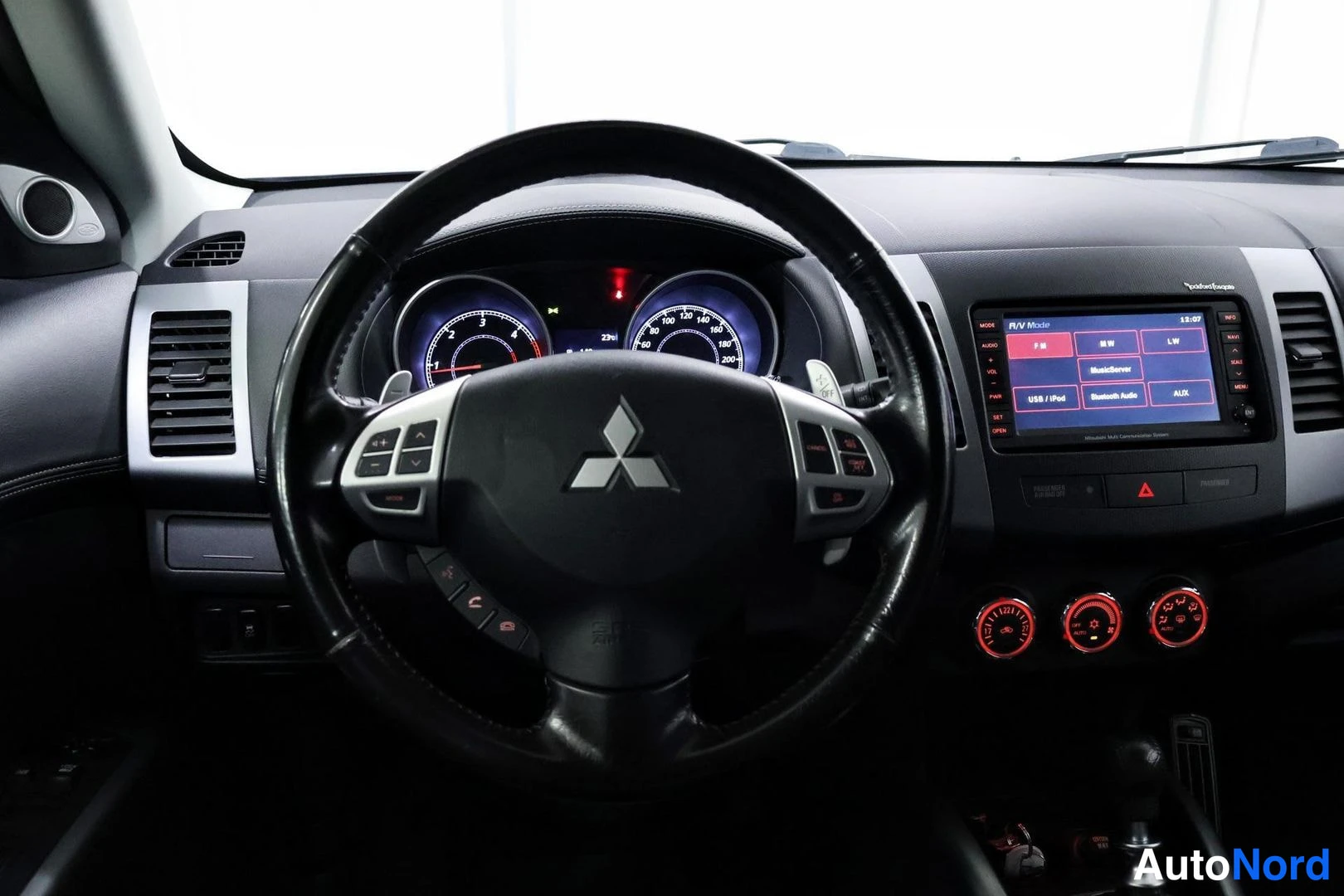 Yksityiskohta: Lähikuva ohjauspyörästä, jossa on Mitsubishi-logo, äänentoiston ja puhelinkytkintä sekä mittariyksikkö taustalla. Keskikonsolissa on GPS-navigointilaite ja puna