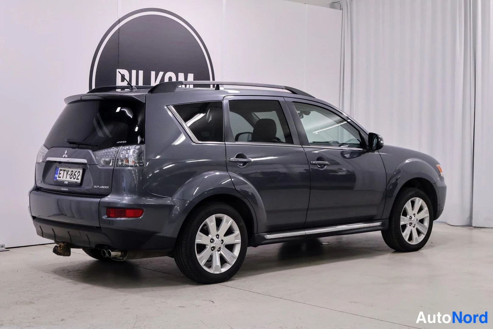 Takaviisto: Takakulma-kuva tummanharmaasta Mitsubishi Outlander -autoista, jolla on kattoharjat ja hopeanväriset vanteet. Rekisterikilpi. Yksi takavaloista, tavaratilaa kat