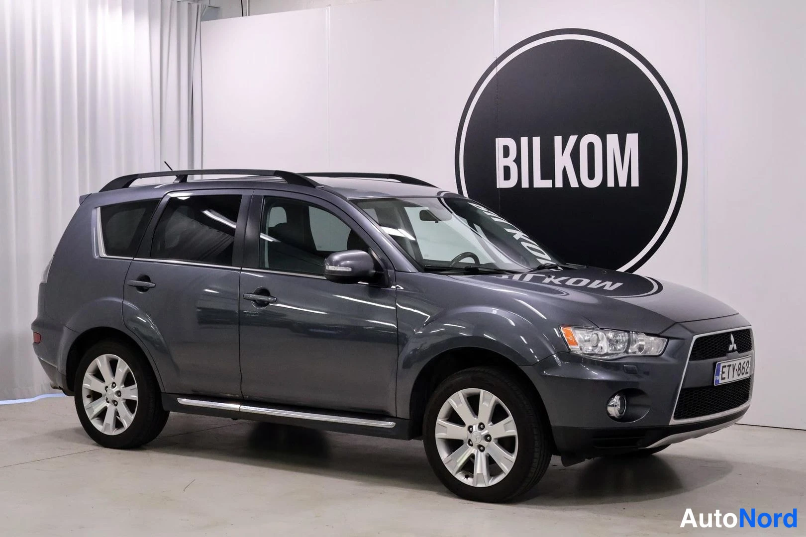 Etuviisto, nokka oikealle: Edessä kulma-kuva tummanharmaasta Mitsubishi Outlanderista, jossa on etusäleikkö ja valot sekä kattoharjat. Auton rekisterikilpi on.