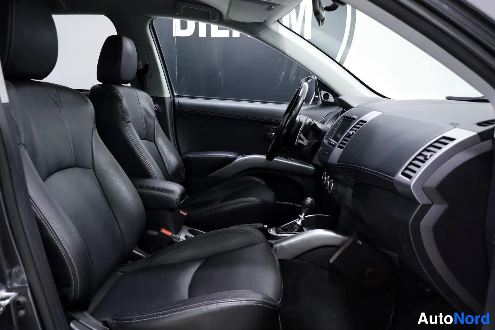Etuistuimet: Black leather front seats with silver trim accents, manual gear lever, and dashboard visible from passenger door opening