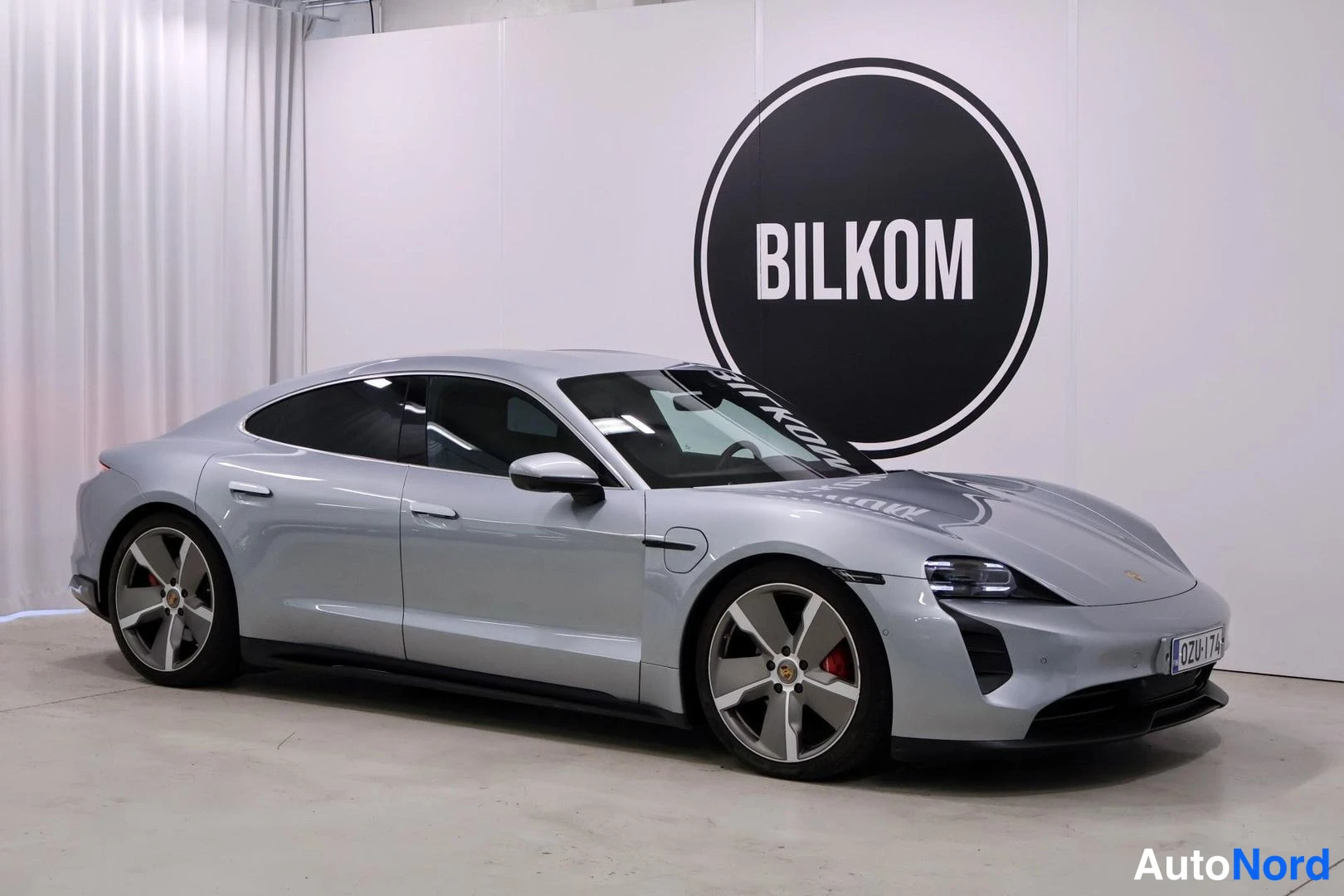 Etuviisto, nokka oikealle: Etukolmionäkymä hopeisesta Porsche Taycanista sisätiloissa; etuveto ja logo näkyvät, BILKOM-logo taustalla