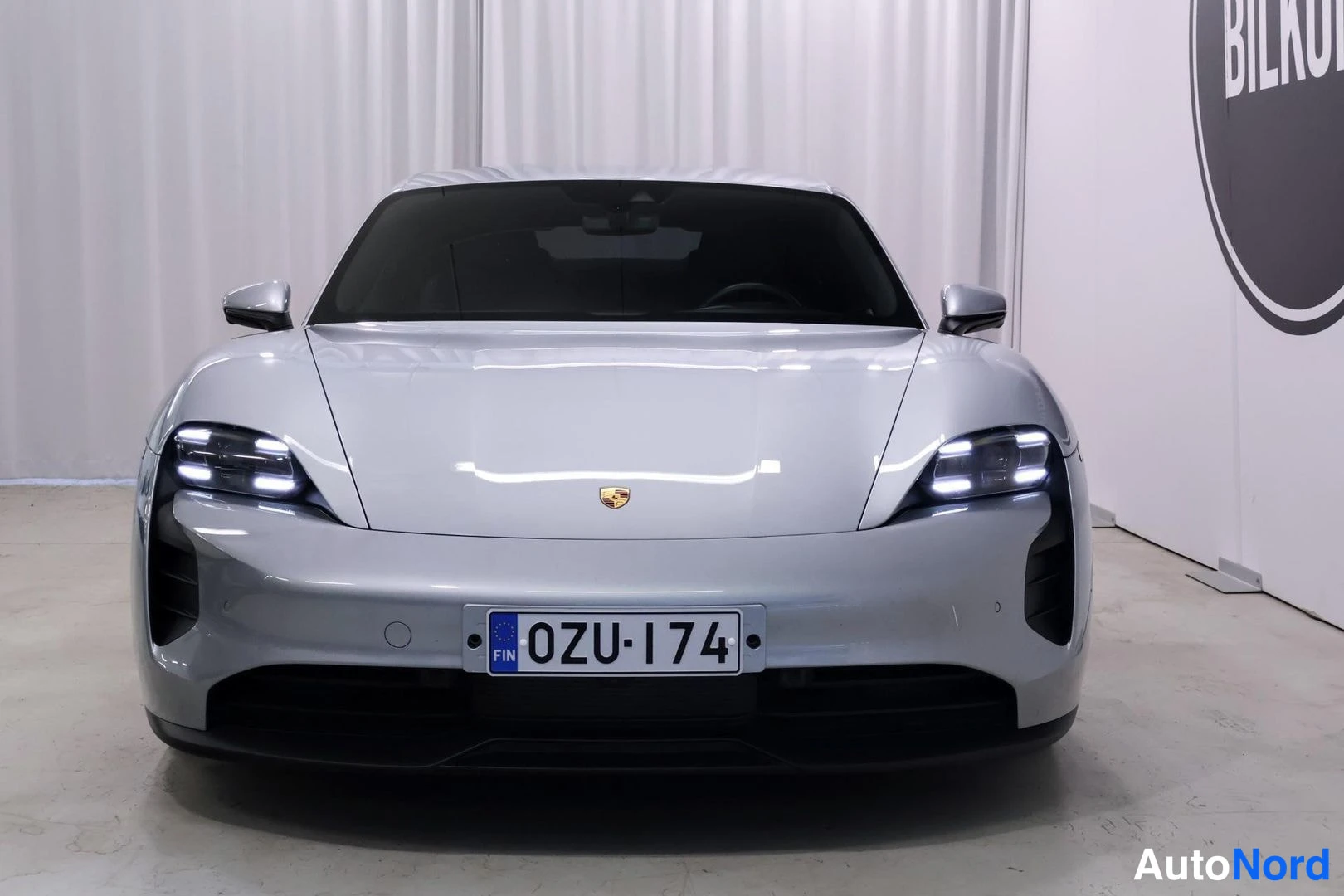 Edestä: Edestä otettu kuva hopeisesta Porsche Taycanista; LED-valot päällä ja suomalainen rekisterinumero