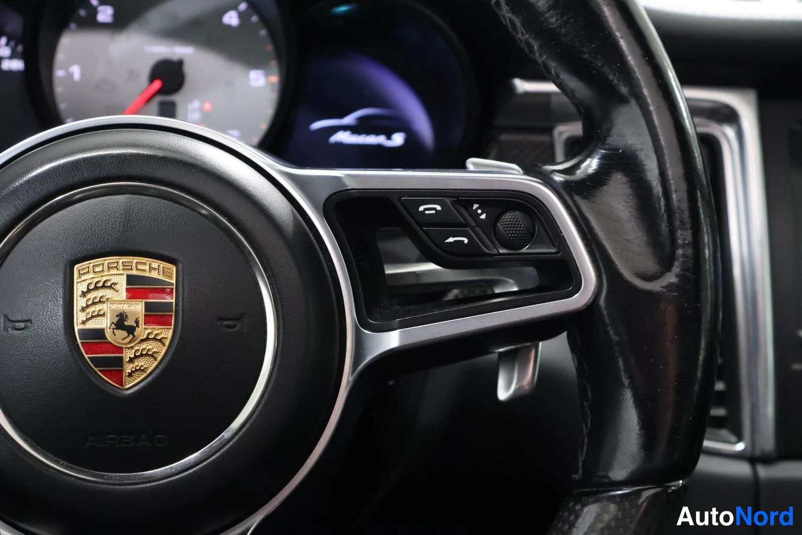 Muu: Ohjauspyörän oikea puolisko Porsche-vaakunalla. Säiliöllä painikkeet puhelin- ja valikkomerkinnöin, taustalla väripöydän näyttö jossa teksti Macan S.