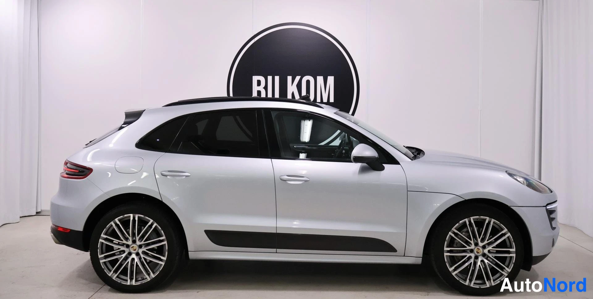 Sivulta: Silver Porsche Macan shown from the left side with a white wall and a round BILKOM logo visible behind it