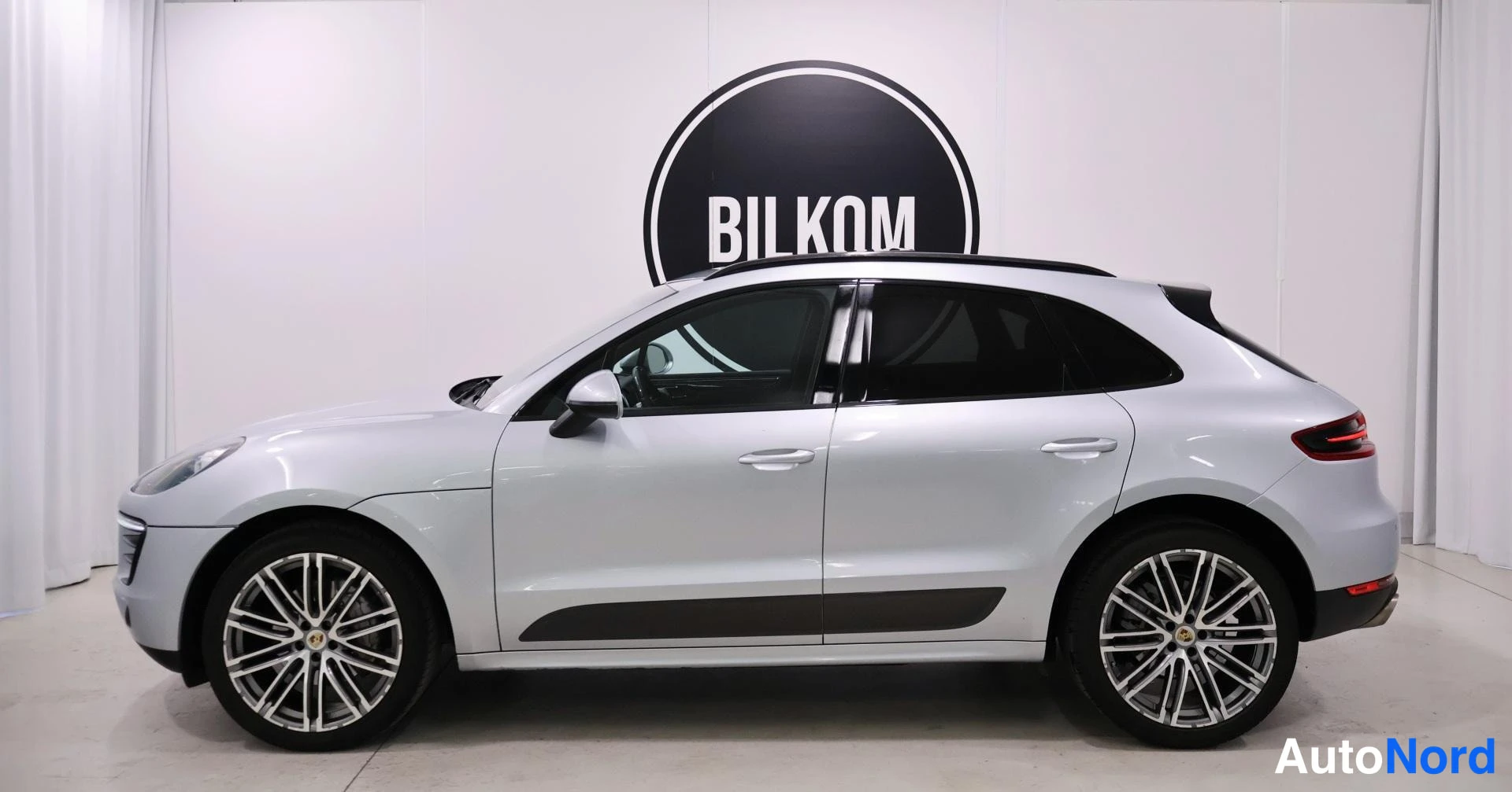 Sivulta: Silver Porsche Macan shown from the right side against a white wall with a black BILKOM sign