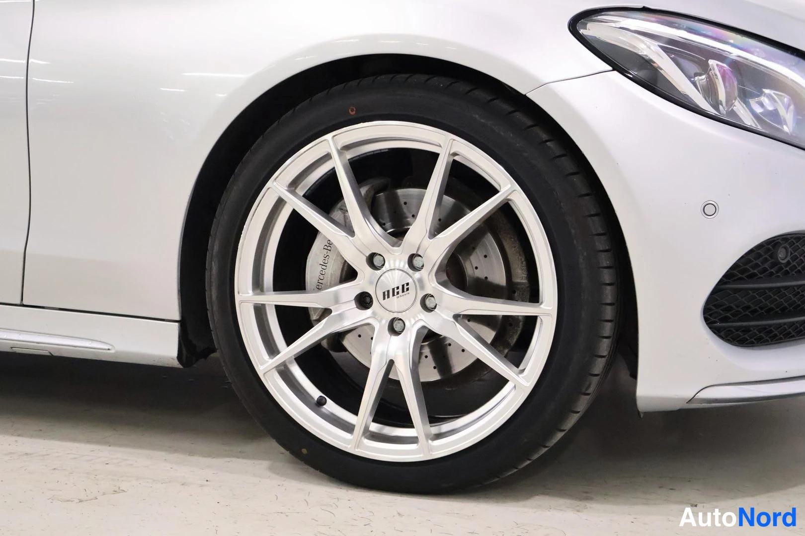 Yksityiskohta: Close-up of white Mercedes-Benz front left wheel showing silver multi-spoke NIB alloy rim and brake caliper with 'Mercedes-AMG' logo visible.