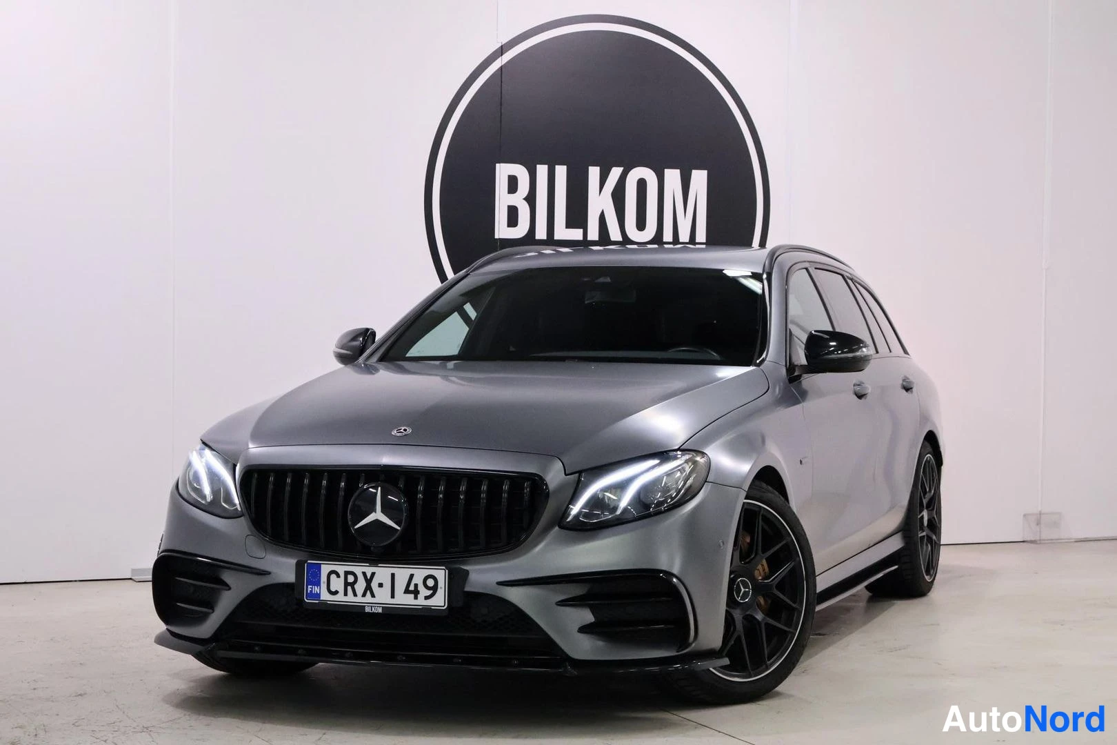 Etuviisto, nokka vasemmalle: Etunäkuvassa näkyy harmaa, mattapintainen Mercedes-Benz-vaunu. Seisoo valkoisen seinän edessä, jossa on musta BILKOM-logo. Autolla on suuri musta etusä