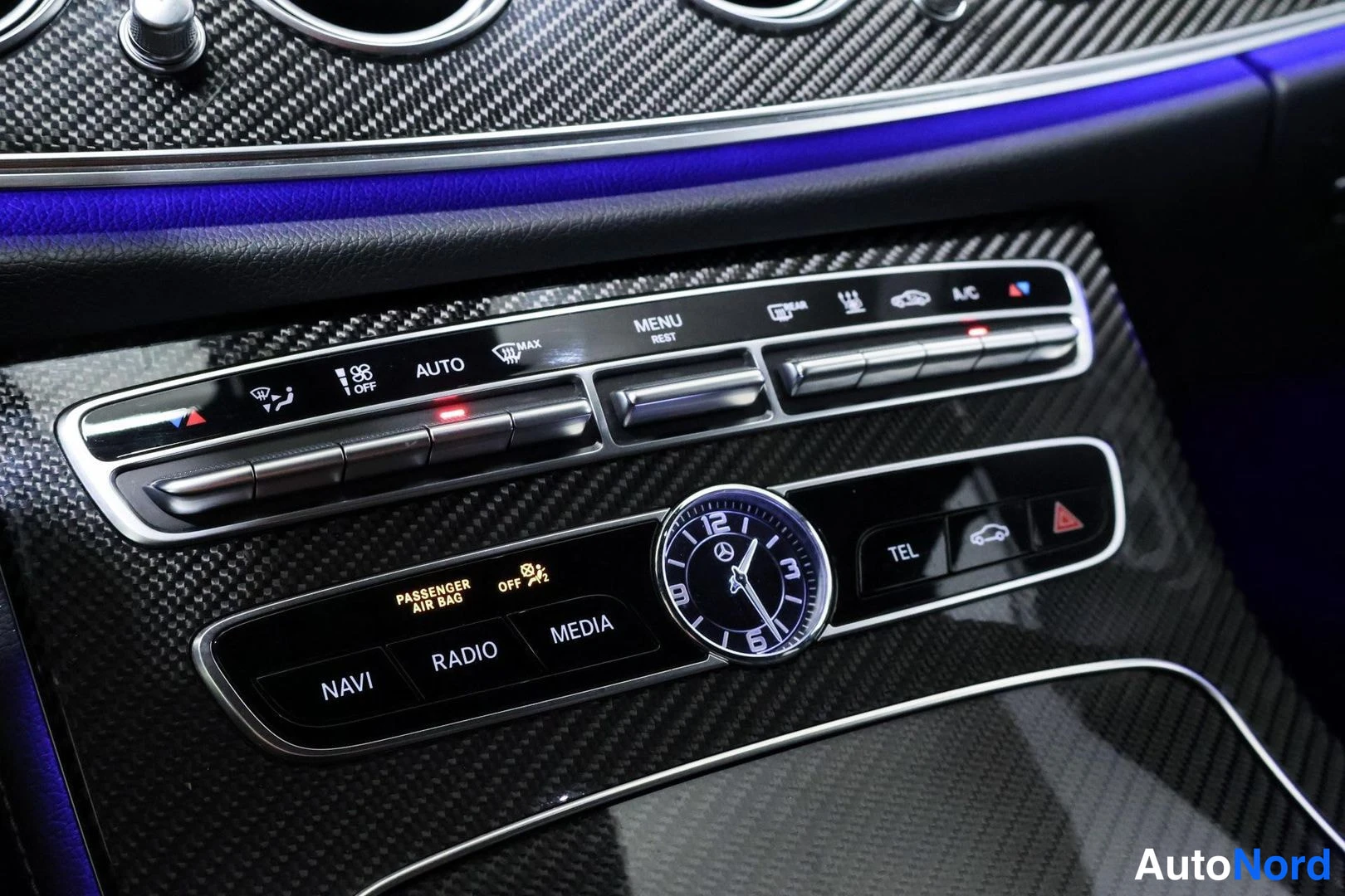 Sivulta: Keskikonsolin hallintapaneeli lähikuva: analoginen kello, ilmastoinnin säädöt (AUTO, MENU, A/C) sekä NAVI-, RADIO-, MEDIA- ja TEL-painikkeet.