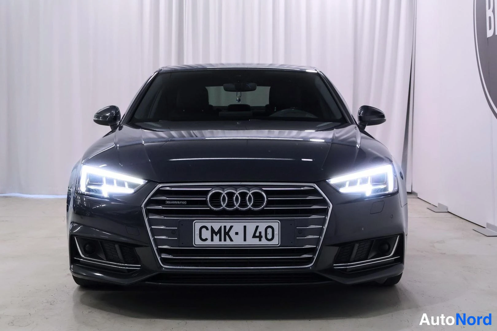 Edestä: Suora kuva mustasta Audi A4:n etuosasta valkoisessa tilassa, näkyvissä ruudukkolattio ja valot.