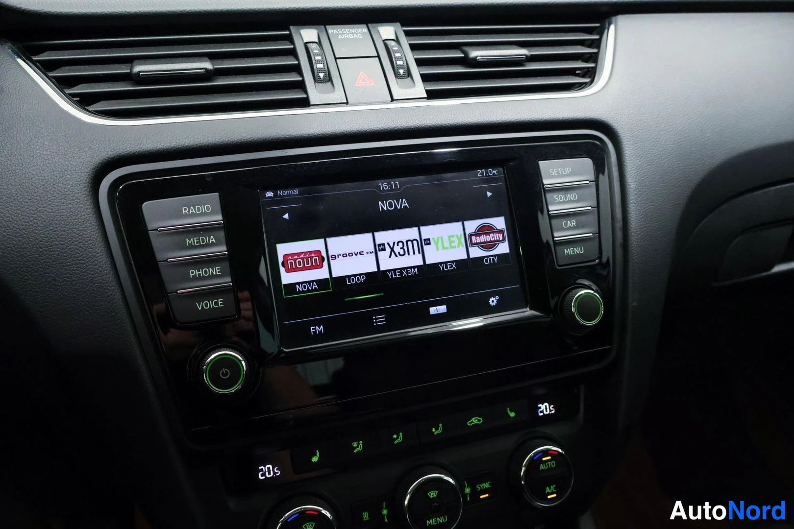 Yksityiskohta: Kojelaudan infotainment-yksikkö radiotoiminnossa, näytöllä NOVA-kanava ja radiolistat, ympärillä RADIO/MEDIA/PHONE/VOICE- ja SETUP/SOUND/CAR/MENU-napit sekä kak