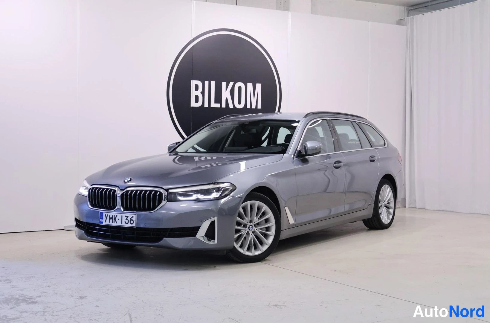 Etuviisto, nokka vasemmalle: BMW 5 -farmari harmaana etukulmasta sisätiloissa, BILKOM-taustataulu.