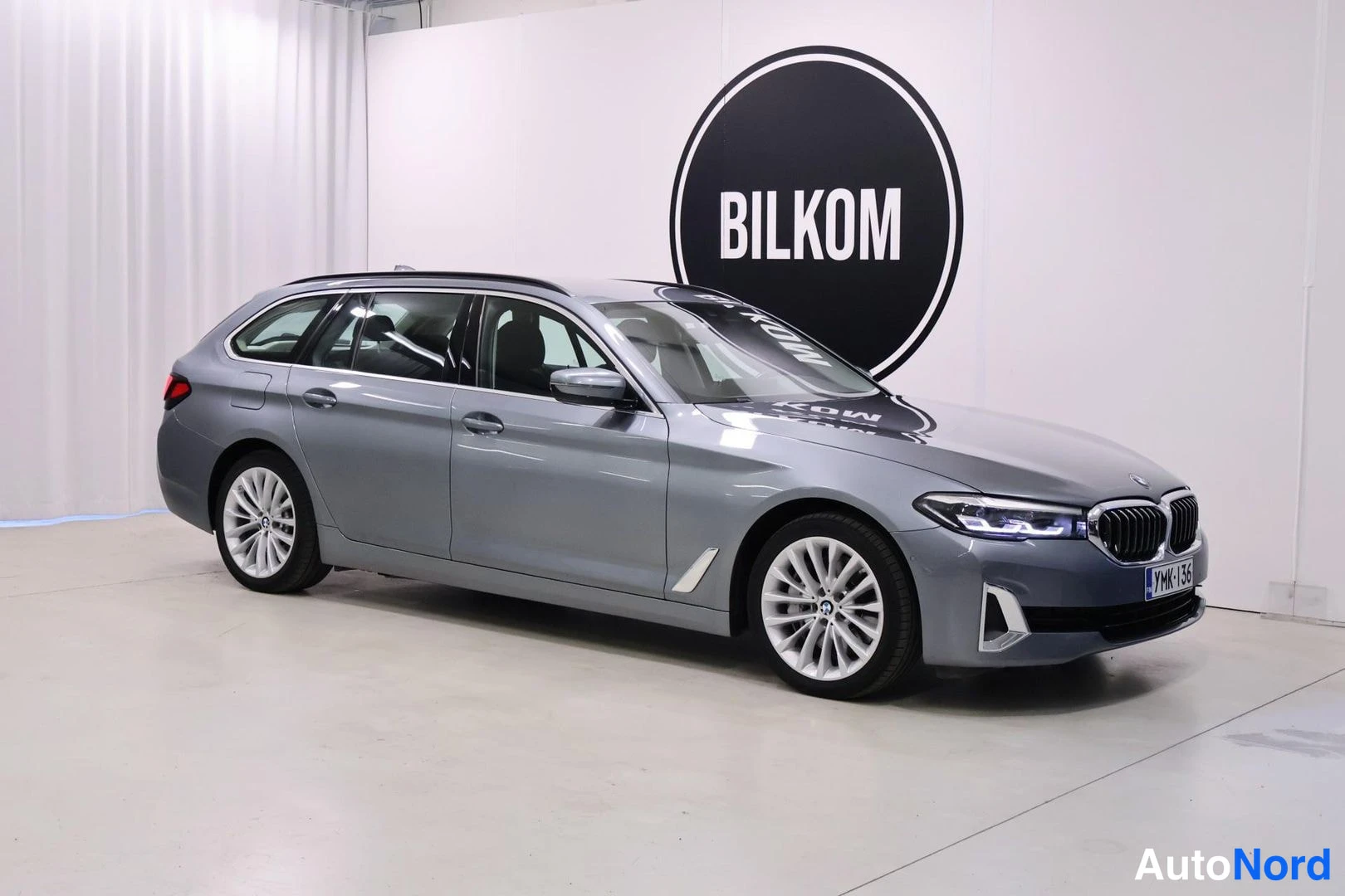 Etuviisto, nokka oikealle: BMW 5 -farmari harmaana etukulmasta sisätiloissa, BILKOM-taustataulu.