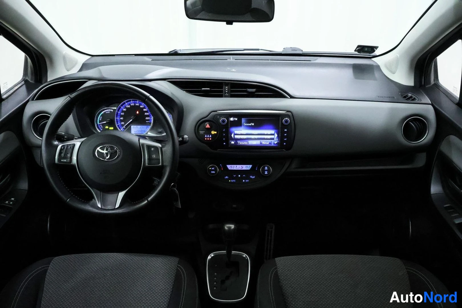 Sivulta: Auton ohjaamo: Toyota-merkkinen ohjauspyörä, mittaristo, kosketusnäyttöradio ja automaattivaihteen valitsin keskikonsolissa.