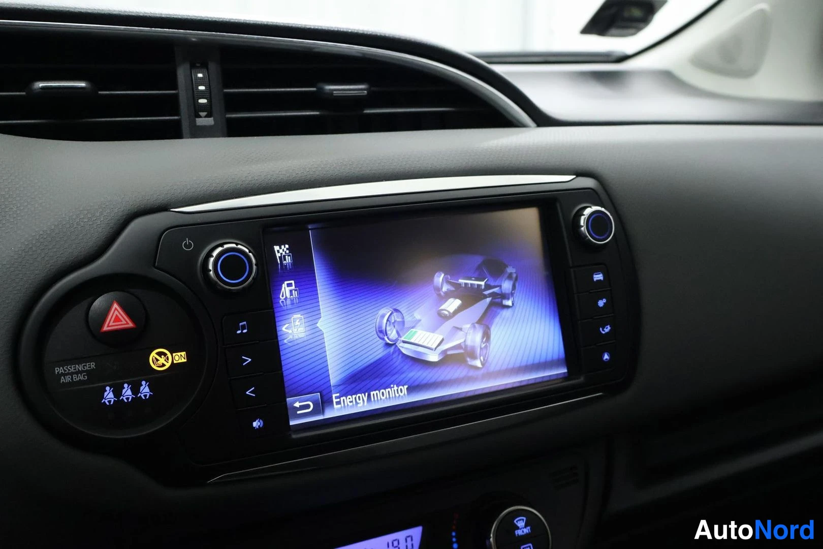 Ohjaamo: Close-up of the center console infotainment screen displaying an 'Energy monitor' graphic showing the hybrid powertrain layout. The hazard light button and pass