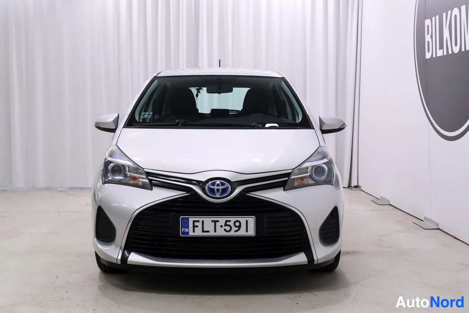 Edestä: Toyota Yaris 2015 kuvattuna suoraan edestä: etusäleikkö Toyota-merkeillä, sumuvalojen aukot ja rekisterikilpi, vaaleanhopea kori vaaleaa verhoa vasten.