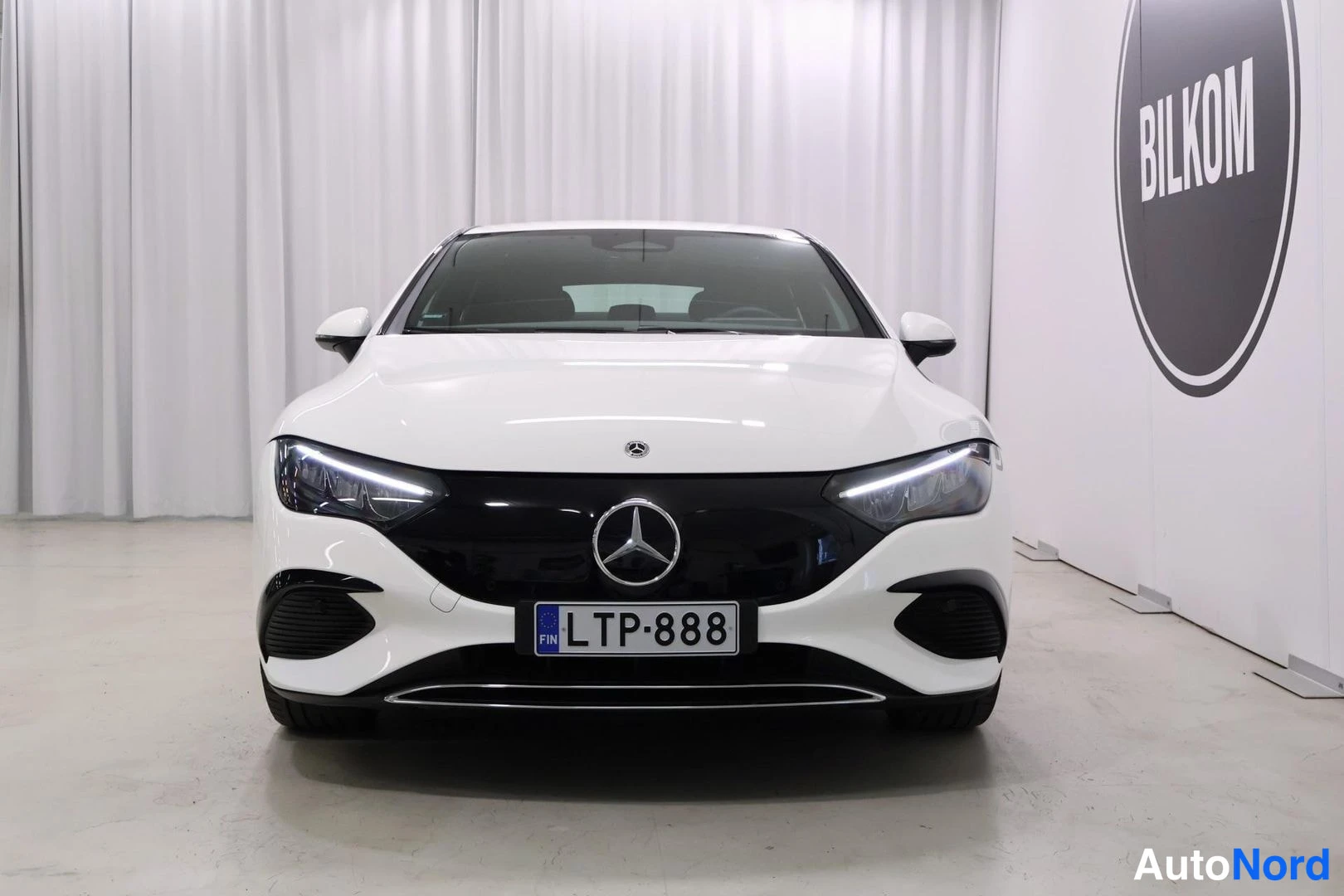 Edestä: Mercedes-Benz EQE -sedanin keula suoraan edestäpäin, valkoinen korimalli, musta maski ja Mercedes-tähti, rekisteritunnus.