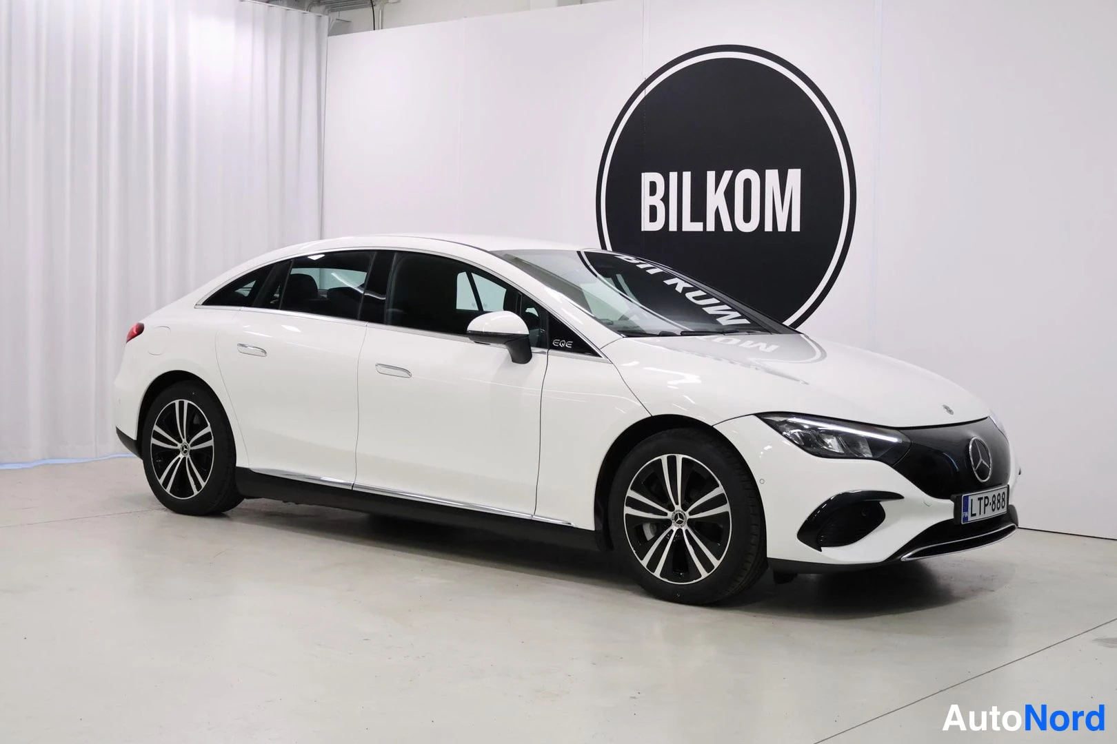 Etuviisto, nokka oikealle: Valkoinen Mercedes-Benz EQE -sedan kulmakuva edestä oikealta, paneeliovi ja mustat kevytmetallivanteet, rekisterikilpi.