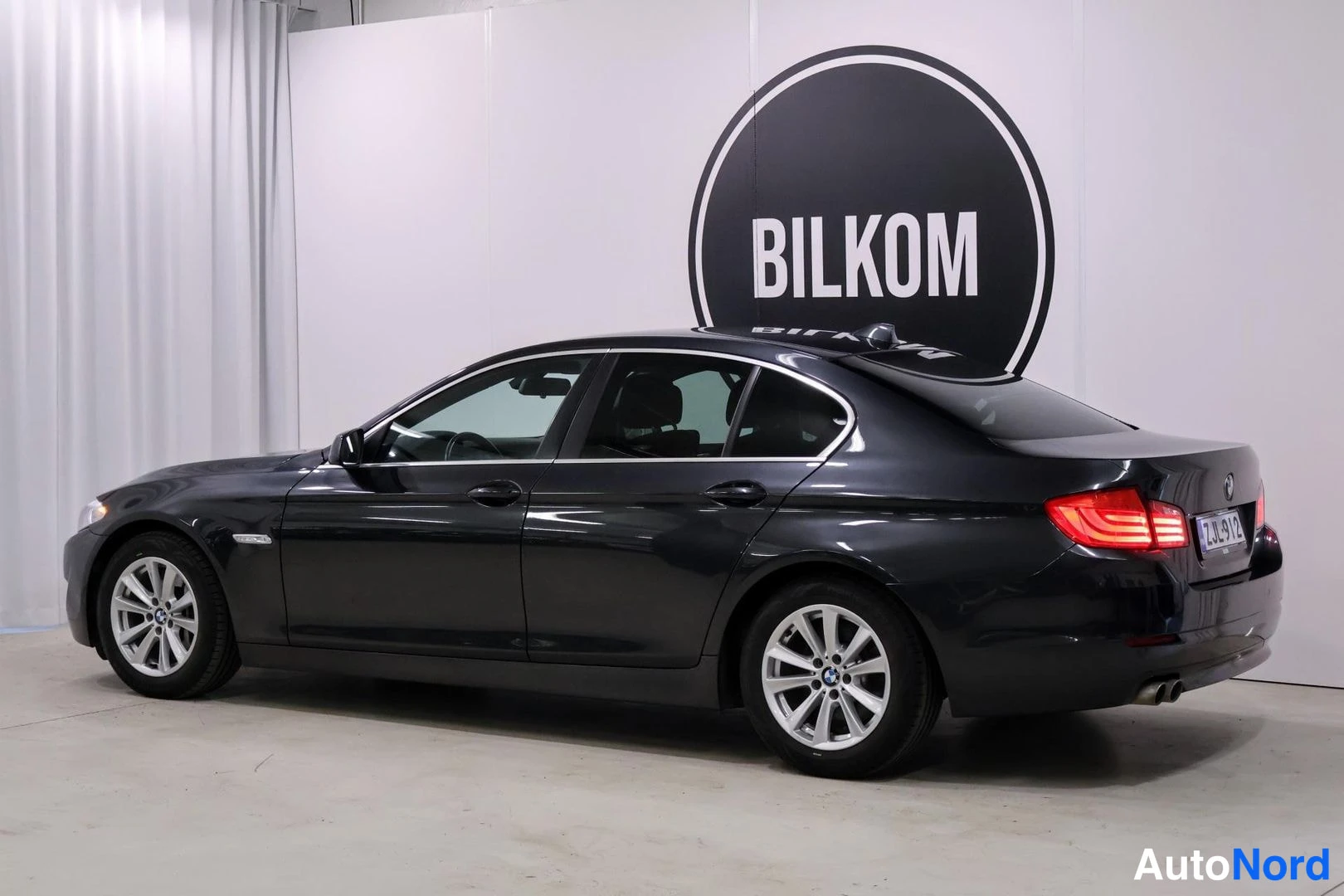 Takaviisto: Mustan harmaan BMW-sedanin takakolmivelanäkymä sisätiloissa, rekisteritunnus.