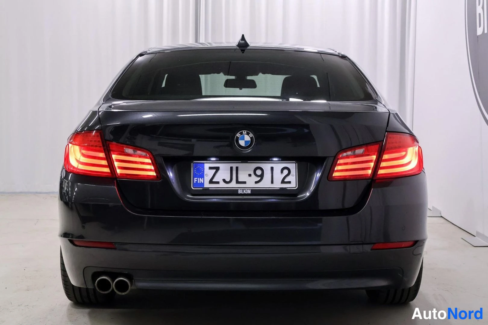 Takaa: Mustan harmaan BMW-sedanin takaosan suoranäkymä, rekisteritunnus ja kaksoispakoputket.