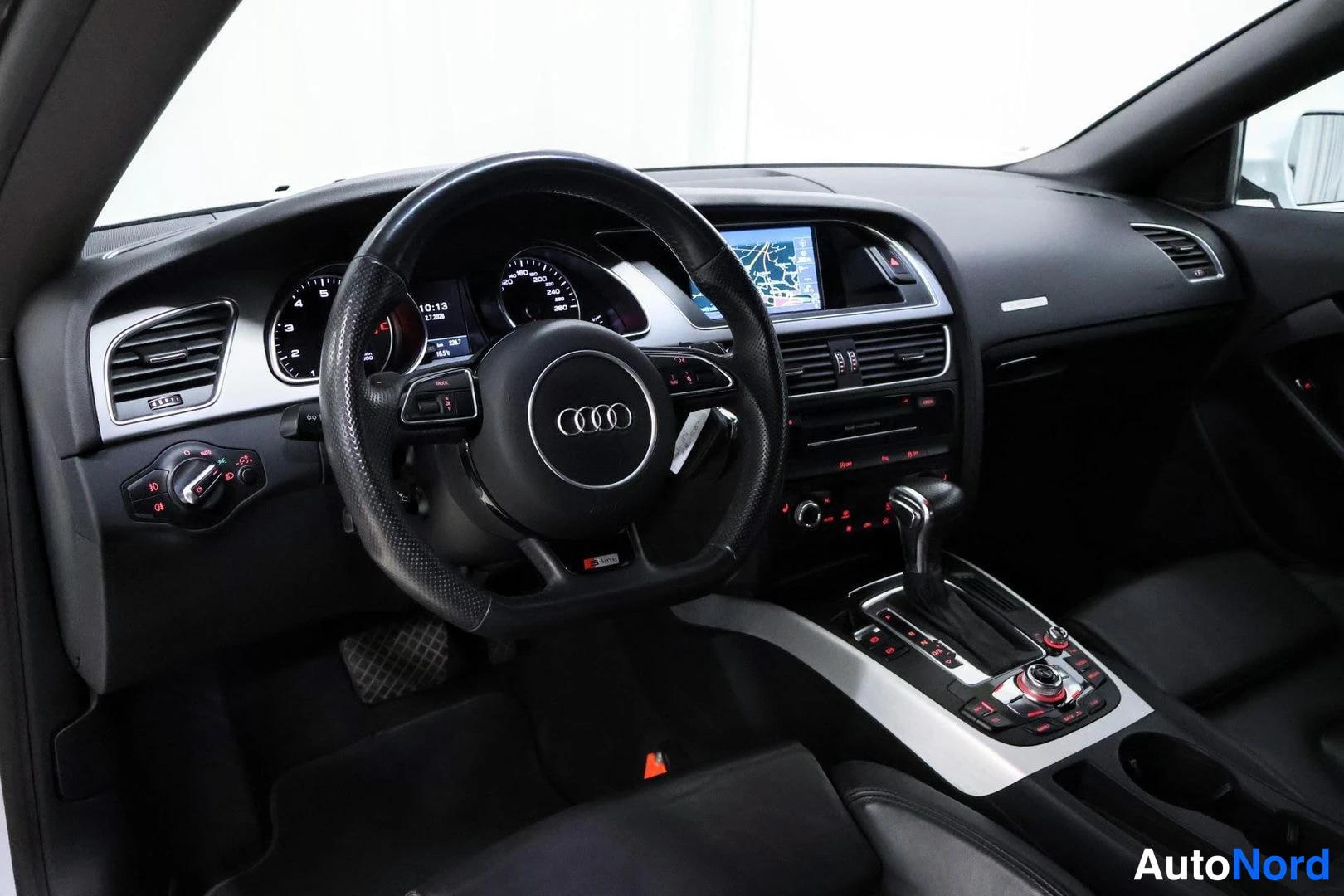 Ohjaamo: Driver's perspective shot featuring a leather-wrapped multifunction steering wheel with paddle shifters, instrument cluster, and a navigation display screen mou