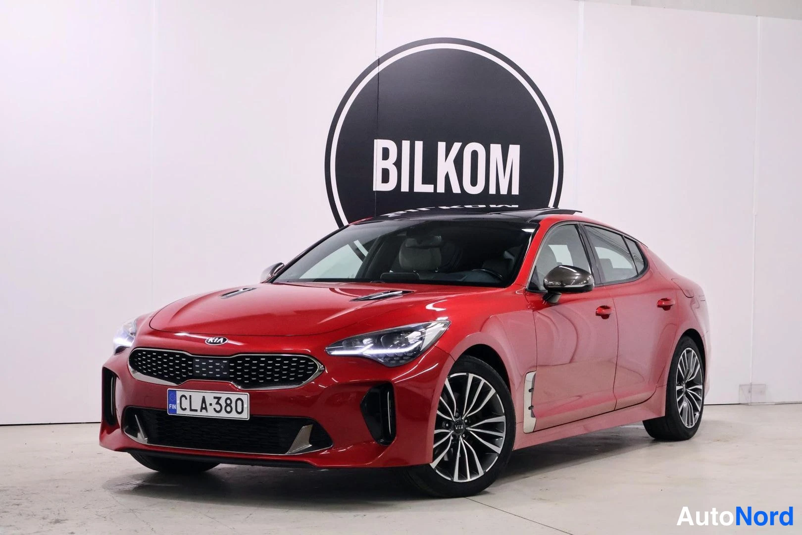 Etuviisto, nokka vasemmalle: Punainen Kia Stinger -henkilöauto etukulmasta sisätiloissa, kilpi.