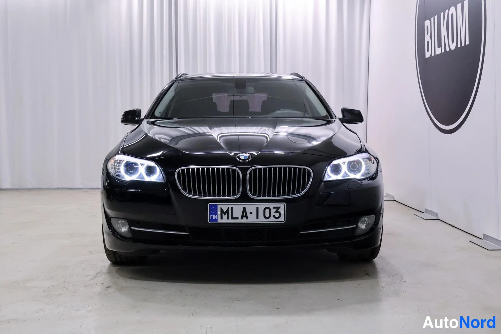 Edestä: Mustan henkilöauton etunäkymä suoraan edestä, BMW-merkki, etuvalot ja rekisteritunnus.