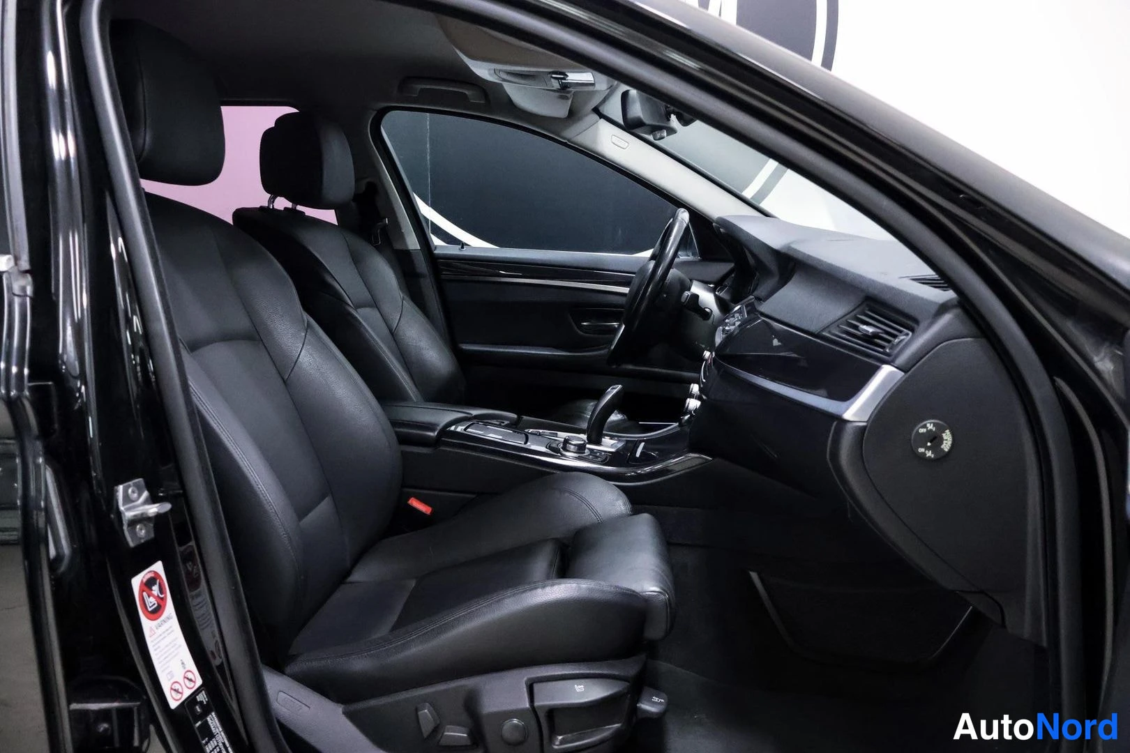 Etuistuimet: Driver side interior view of black leather seats. Dashboard and center console are in frame showing a start-stop engine button and iDrive control knob.