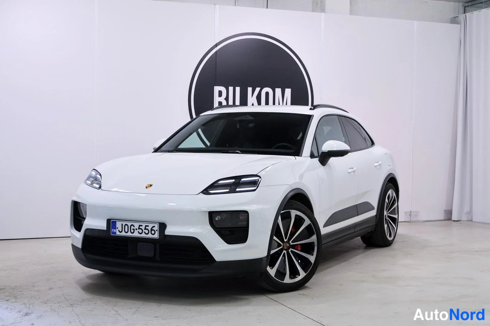 Etuviisto, nokka vasemmalle: Valkoinen Porsche Macan kolmeneljäsosan etunäkymässä keula vasemmalla, rekisteritunnus, kuvattu sisätilassa valkoista seinää ja BILKOM-logoa vasten.