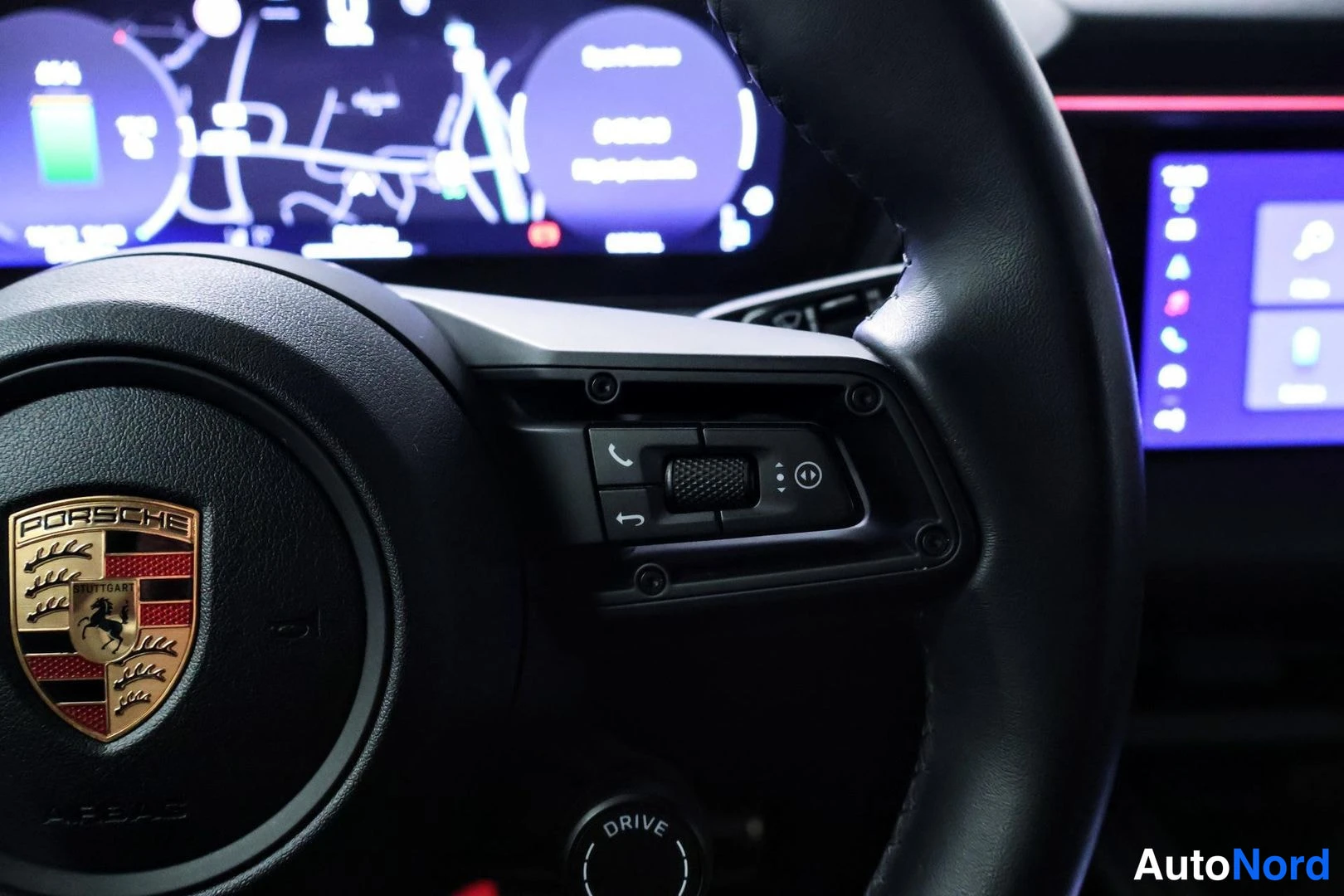Yksityiskohta: Close-up of the right side of a black steering wheel showing Porsche crest, control buttons for phone calls and settings, and part of the central digital displa
