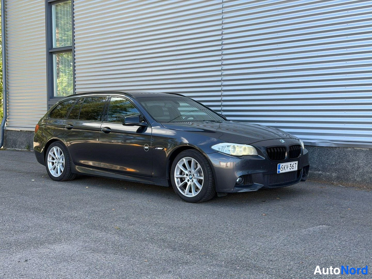 Etuviisto, nokka vasemmalle: Etu-oikea viistokuva tummanharmaasta BMW 5-sarjan farmariautosta. Auton etuosa ja oikea kylki näkyvät kuvassa.