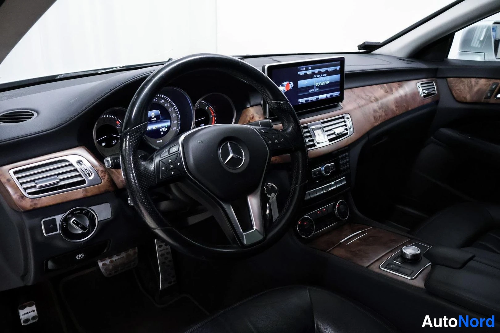 Edestä: Ohjaamon sisäkuva: musta Mercedes-monitoimiohjauspyörä, analoginen mittaristo, kojelautanäyttö ja puukuvioinen keskikonsoli mustilla nahkapinnoilla.