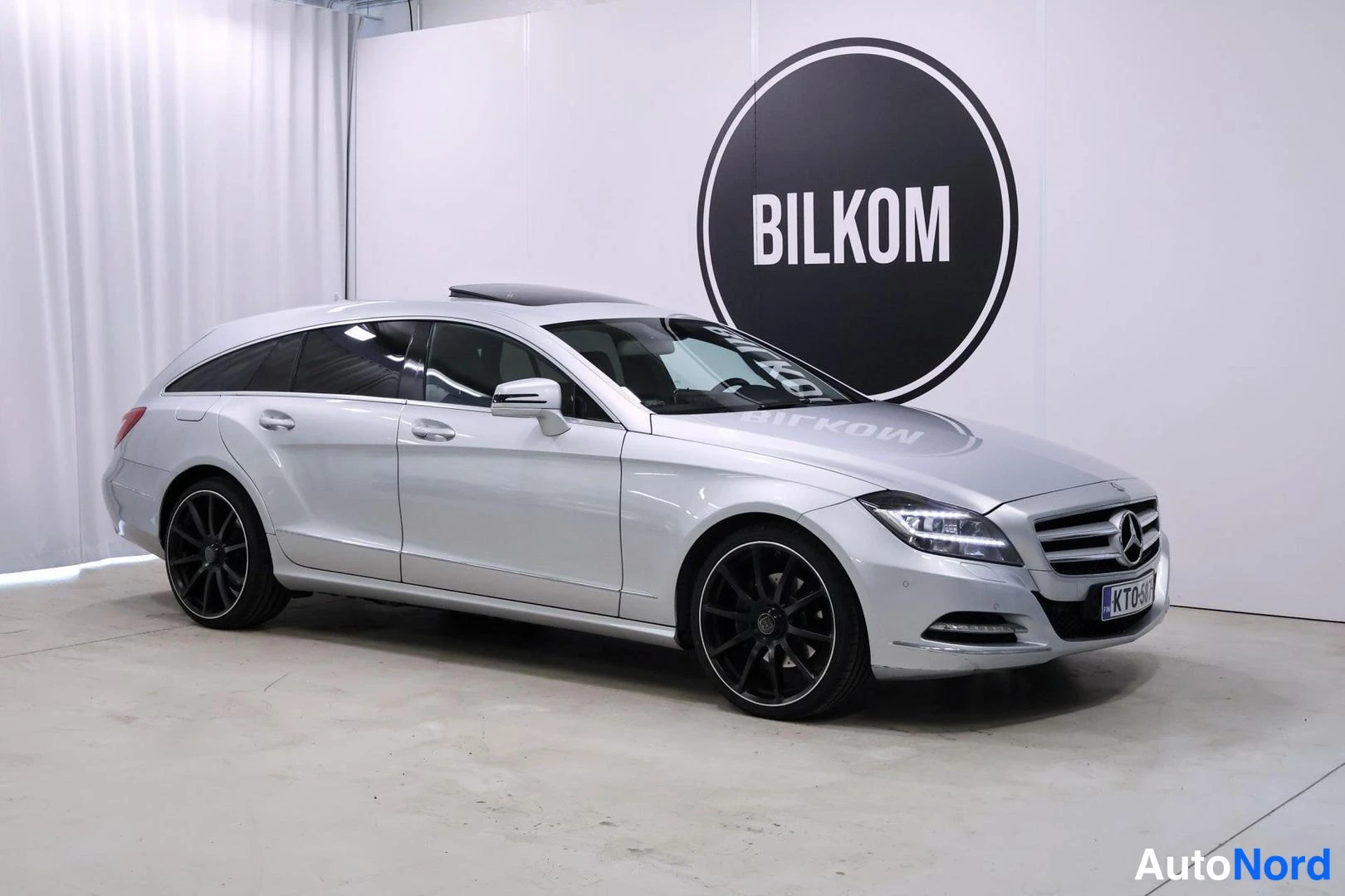 Etuviisto, nokka oikealle: Valkoinen Mercedes-Benz CLS -farmari seisoo sisätiloissa. Takakatto on avattava. Mustat kevytmetallivanteet näkyvät. Taustalla valkoisella seinällä musta
