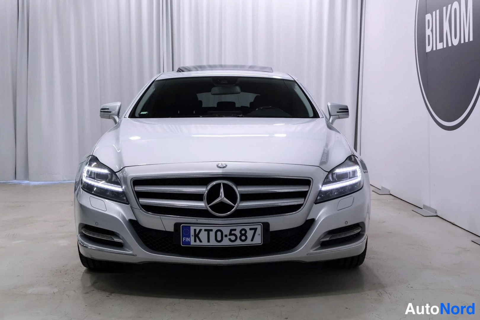 Edestä: Etukuvassa valkoinen Mercedes-Benz -auto. LED-ajovalot ja etusäleikkö ovat selkeästi näkyvissä. Eturekisterikilpi lukee. Seinällä oikealla BILKOM-logon osa.