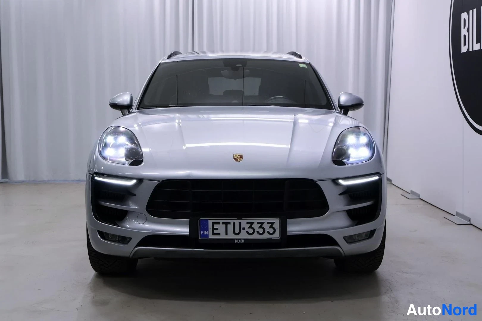 Edestä: Auton etunäkymä, hopea Porsche Macan, rekisterikilpi, etuvalot päällä.