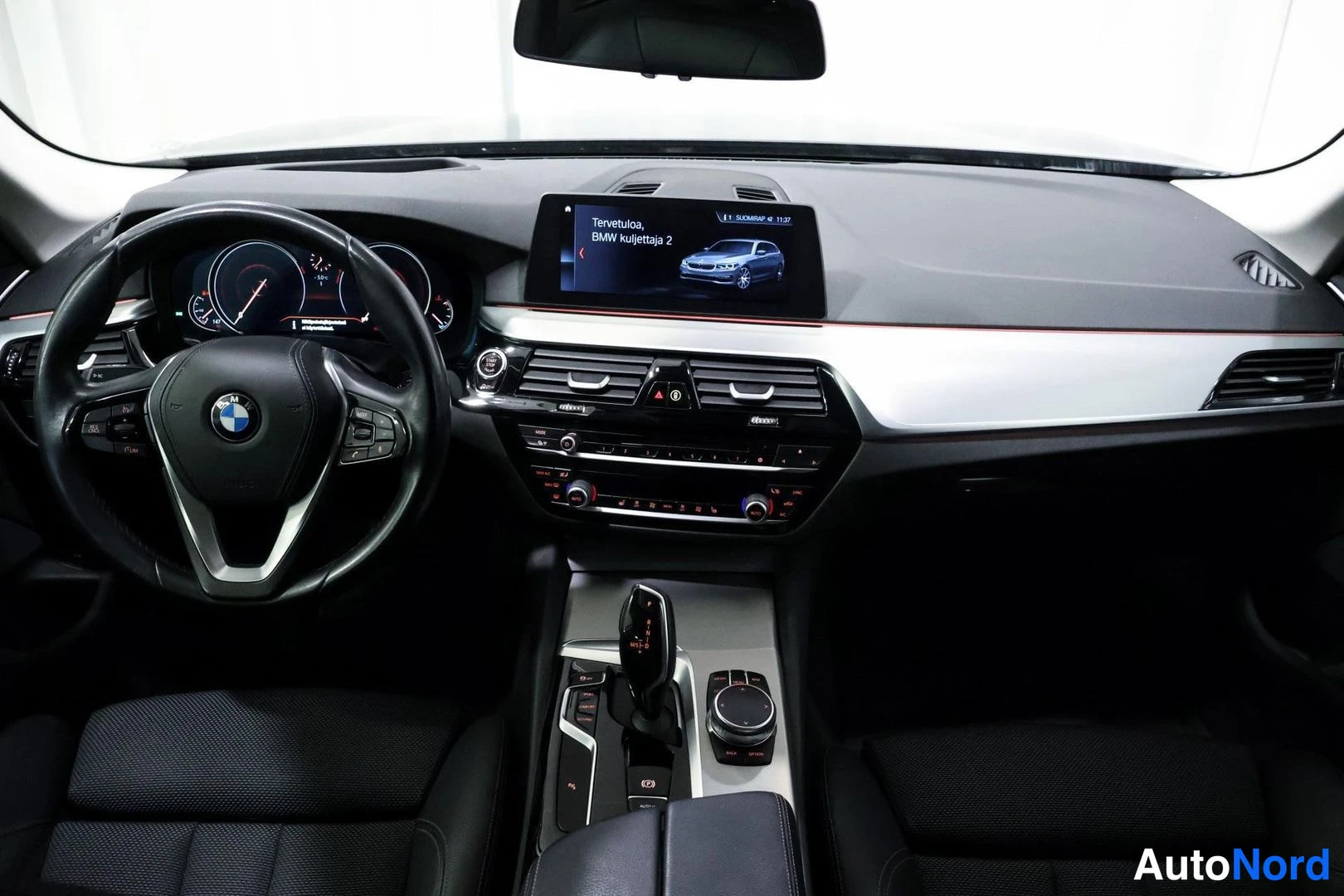 Muu: Ohjaamo suoraan edestä: BMW-ratti, mittaristo, keskitoiminnot ja näyttö tekstillä 'Tervetuloa, BMW kuljettaja 2'.
