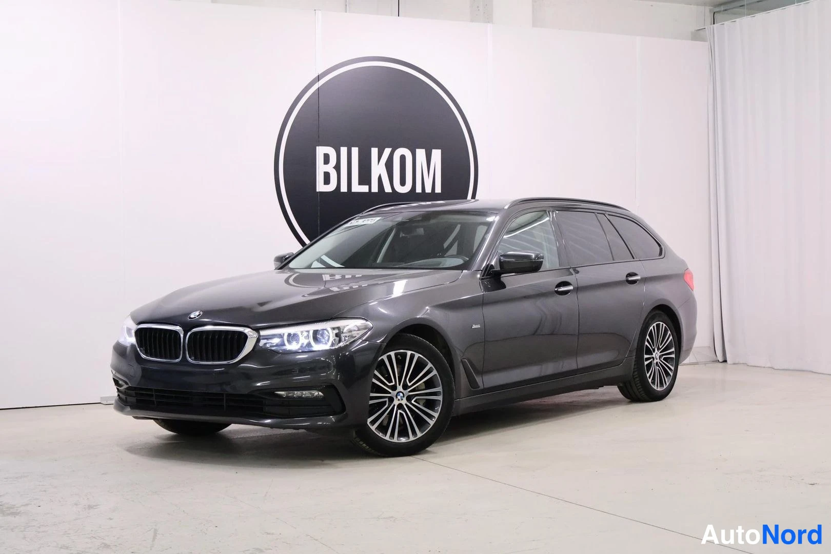 Etuviisto, nokka vasemmalle: Tummanharmaa BMW 530 Station Wagon seisoo sisätiloissa valkoisten seinien edessä. Etuvalot päällä. Bilkom-logo seinässä.